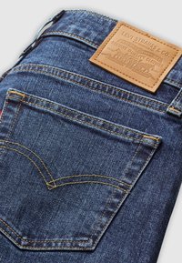 Jeans en denim bleu foncé avec une étiquette en cuir, des coutures visibles et un design de poche arrière classique avec une courbe distinctive.