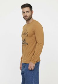 Langarm, mosterdkleurig shirt met een grafische print van een eland en bomen. Gemaakt van zachte stof, met een ronde halslijn en een relaxte pasvorm.
