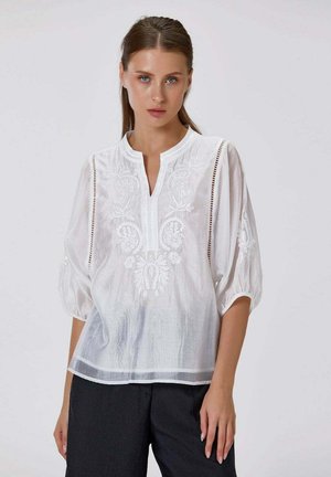 Femme aux cheveux bruns et raides portant un chemisier blanc brodé avec manches ballon 3/4 et un pantalon noir sur fond uni.