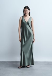Femeie purtând o rochie maxi din satin verde măsliniu, fără mâneci și cu decolteu adânc în V, și papuci negri, stând pe un fundal simplu gri deschis.