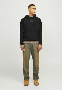 Sudadera con capucha negra y bolsillo frontal, combinada con pantalones beige y botines marrones. Presenta cordones y una etiqueta decorativa en el pecho.