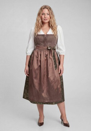 Frau mit langen welligen blonden Haaren, die ein braunes traditionelles Kleid mit weißen Puffärmeln und passenden hohen Absätzen trägt, steht vor grauem Hintergrund.