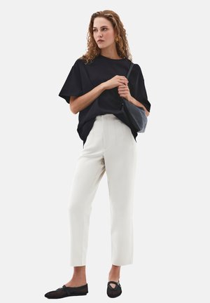 Femme portant une chemise noire oversize, un pantalon blanc ajusté, des ballerines noires, tenant un sac à bandoulière noir, debout devant un fond uni.