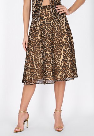 Kobieta ubraną w brązową i czarną midi spódnicę w panterkę, zestawioną z złotymi sandałami na wysokim obcasie z paskiem wokół kostki, stojąca z jedną ręką na biodrze.