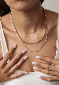 Warstwowe naszyjniki w różowym złocie z projektami ogniw, spinaczy papierowych i delikatnych łańcuchów, prezentowane na skórze. Widać długie, gładkie, jasnoróżowe paznokcie.