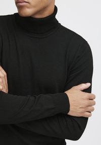 Schwarzer gerippter Rollkragenpullover mit langen Ärmeln. Weicher Stoff, taillierte Passform und ein enger Kragen. Arme vor der Brust verschränkt.