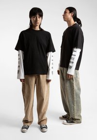 Zwart T-shirt met korte mouwen over een wit shirt met lange mouwen, gecombineerd met beige corduroy en lichtblauwe spijkerbroek. Zwarte sneakers.