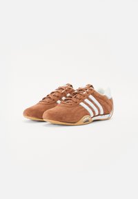 Barna velúr sportcipők fehér csíkokkal, lekerekített orral, fűzős kialakítással és gumi talppal, az oldalon "adidas" logóval.