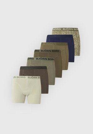 Muskulöser Mann steht barfuß mit verschränkten Armen, trägt dunkle Boxershorts vor schlichtem hellem Hintergrund.