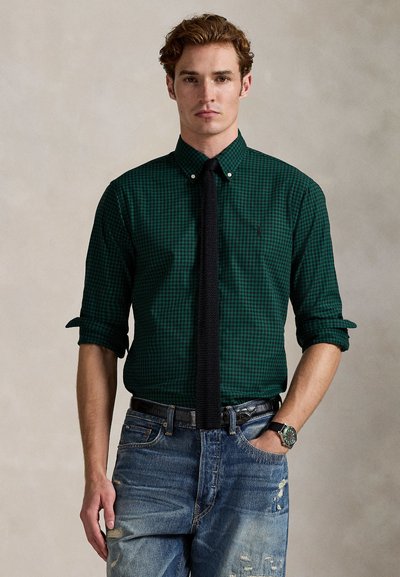 Young man wearing a dark green checkered shirt with rolled sleeves, black tie, worn blue jeans, black belt, and wristwatch, standing against plain background.