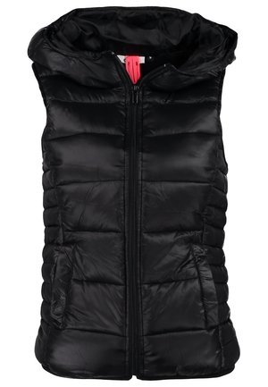 Jeune personne portant une doudoune noire sans manches sur une chemise blanche à manches longues, jean noir, baskets bleu marine, portant un sac à dos noir sur une épaule.