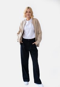 Beżowa bluza z zamkiem, biała koszulka i czarne szerokie spodnie z kieszeniami. Model stoi w swobodnej pozie na białym tle.