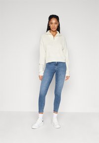 Suéter de punto blanco con cuello con cremallera, combinado con jeans ajustados azules y zapatillas blancas. El conjunto presenta un ajuste relajado y una tela texturizada.