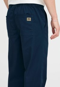 Marineblauwe broek met een gladde textuur, elastische tailleband en twee achterzakken. Bevat een klein label op de rechterzak.