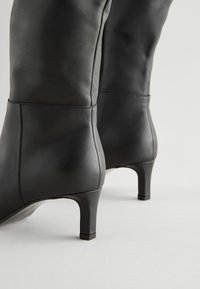 Bottes mi-mollet en cuir noir avec talons bas et fins sur un fond clair uni, montrant les détails de dos et de côté.