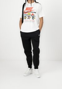 Nike weißes T-Shirt mit buntem grafischen Druck, das Charaktere zeigt, kombiniert mit schwarzen Jogginghosen und weißen Sneakers mit Air-Dämpfung.