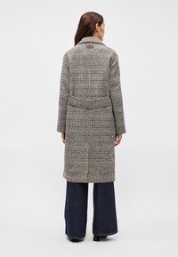 Καρό παλτό σε γκρι, μπεζ και καφέ αποχρώσεις, μεσαίου μήκους, με ζώνη στη μέση, κολάρο με αυτιά και ίσια γραμμή, σε συνδυασμό με φαρδιά τζιν.