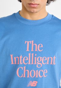 SVětle modré bavlněné tričko s nápisem „The Intelligent Choice“ ve výrazném růžovém písmu a malým bílým logem New Balance ve spodní části.