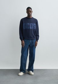 Marinblå sweatshirt med "GCDS"-logotyp, i kombination med löst sittande blå jeans och vita sneakers. Tydlig, modern design och bekväm passform.