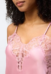 Κοντινό πλάνο μιας γυναίκας που φοράει ροζ σατέν καμισόλ με φλοράλ δαντέλα στο τελείωμα και λεπτές ράντες στους ώμους.