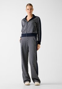 Marineblauw trainingspak met geometrisch patroon, jack met rits, elastische tailleband en zijzakken. Zachte stof, ontspannen pasvorm, gecombineerd met witte schoenen.