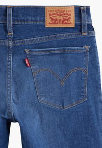 Jeans de mezclilla azul oscuro con un diseño clásico de bolsillo trasero, costuras en contraste y una etiqueta de marca de cuero marrón con detalles grabados.