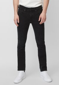 Hombre con pantalones de pana negros ajustados y zapatillas blancas, de pie contra un fondo gris claro liso.