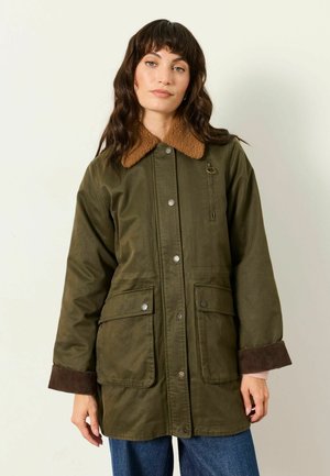 Frau trägt eine olivgrüne Jacke mit braunem Lammfellkragen und großen Fronttaschen, steht vor einem schlichten hellen Hintergrund.