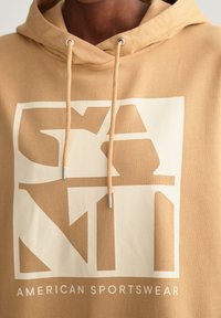 Sweat à capuche beige avec un grand motif blanc comportant des lettres angulaires et le texte "AMERICAN SPORTSWEAR" en bas. Cordon de serrage avec embouts en métal.