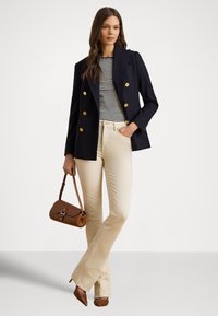 Sieviete tērpusies tumši zilā dubultpogātas žaketes, svītrainas krekla, bēša krāsas biksēs, brūnās augstpapēžu kurpēs un ar brūnu rokassomiņu, stāvot pret vienkārša baltā fona.