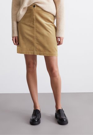 Kobieta ubrana w beżowy sweter i mini spódniczkę ze sztruksu w kolorze brązowym, nosząca czarne wsuwane mokasyny, stoi na szarej podłodze na tle jednolitego tła.