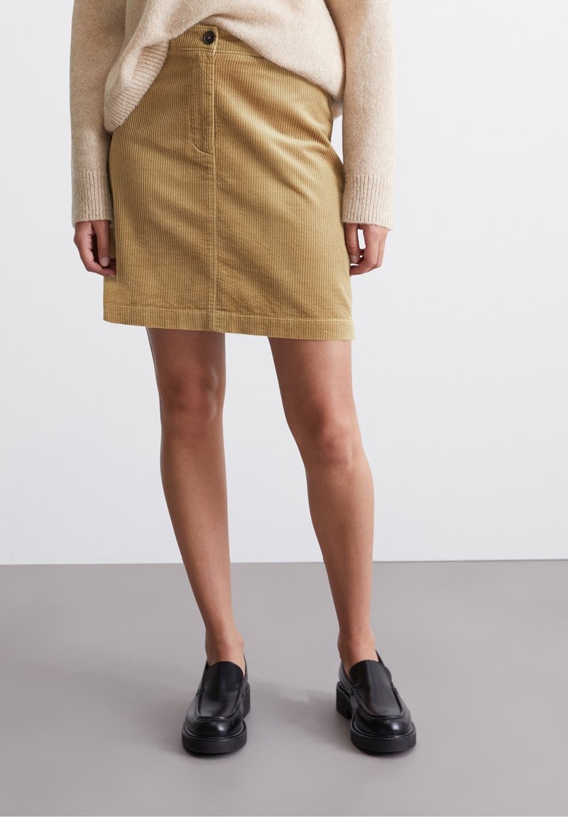 Femme portant un pull beige et une mini-jupe en velours côtelé couleur chameau avec des mocassins noirs sans lacets, debout sur un sol gris devant un fond uni.
