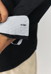 Črna srajca z modro-belim črtastim manšetom, ki izstopa z detajlnim šivom in subtilnim logotipom. Material je videti bombaž, z gladko teksturo.