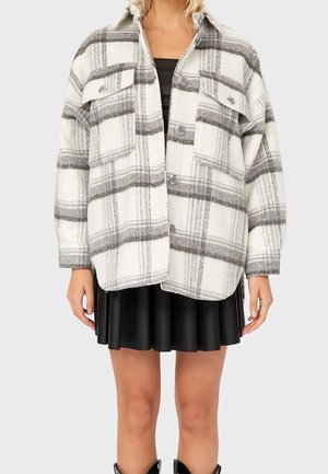 Femme portant une veste boutonnée oversize à carreaux blancs et gris par-dessus une robe noire avec une jupe plissée, visible des épaules aux genoux.