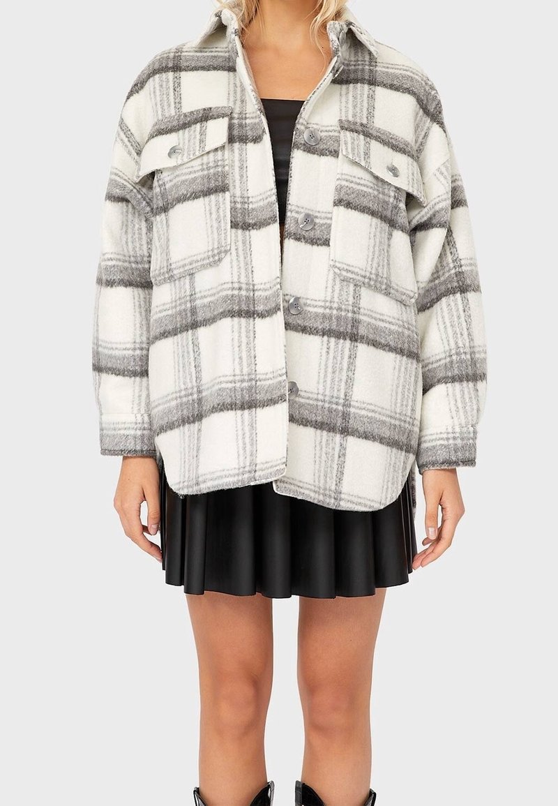 Femme portant une veste boutonnée oversize à carreaux blancs et gris par-dessus une robe noire avec une jupe plissée, visible des épaules aux genoux.