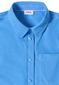 Chemise en velours côtelé bleu clair avec col pointu, boutons sur le devant, poche poitrine et étiquette de marque "Cecil" à l'intérieur du col.