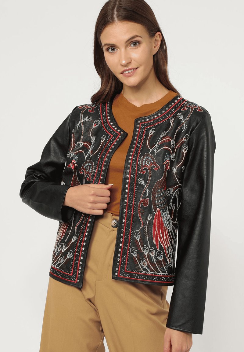 Mujer vistiendo una chaqueta de cuero negra con bordado de pavo real en rojo y blanco, blusa marrón y pantalones beige, de pie contra un fondo liso.