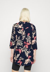 Donna con capelli biondi corti che indossa una blusa blu navy con motivo floreale e farfalle a maniche tre quarti e pantaloni shorts neri, vista da dietro.
