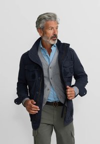 Chaqueta navy con múltiples bolsillos, suéter beige y camisa azul clara, combinados con pantalones cargo oliva. Presenta un cuello alzado y cremallera.