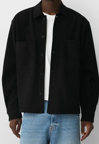 Mężczyzna ubrany w czarną kurtkę zapinaną na guziki z dwoma przednimi kieszeniami, pod którą ma białą koszulę i jasnoniebieskie dżinsy, widoczny od ramion do pasa.