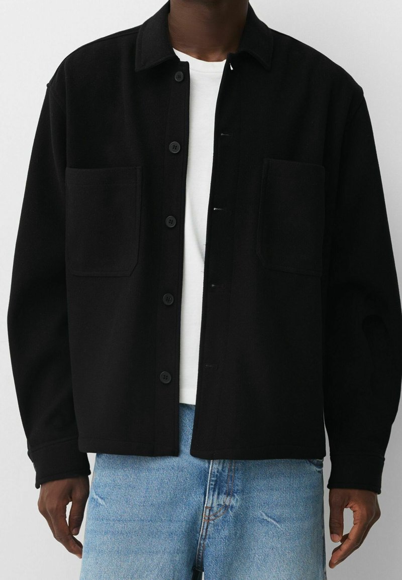 Mężczyzna ubrany w czarną kurtkę zapinaną na guziki z dwoma przednimi kieszeniami, pod którą ma białą koszulę i jasnoniebieskie dżinsy, widoczny od ramion do pasa.