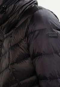 Mustapuffer-takki, jossa on tikattu muotoilu, kiiltävä nailonmateriaali, vetoketjusuljenta ja pieni etiketti hihassa, jossa lukee "Q/S suunniteltu".