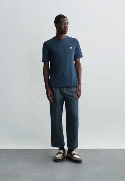 Tricou din bumbac bleumarin cu un mic logo portocaliu, asortat cu pantaloni gri lejeri și pantofi bicolori cu talpă groasă și detalii decorative.
