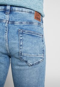 Los jeans de denim azul claro presentan un diseño clásico de cinco bolsillos. La textura es ligeramente desgastada, con una etiqueta de cuero en la parte trasera de la cintura.