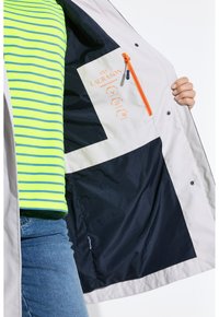 Veste de couleur claire avec un intérieur bleu marine, dotée d'une poche zippée orange et d'un patch logo. Un haut rayé vert vif est superposé en dessous.