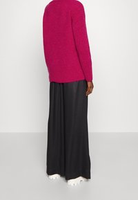 Fuchsiový pletený svetr s volným střihem, žebrovaným lemem a spadlými rameny, v kombinaci s širokými černými kalhotami a bílou obuví.