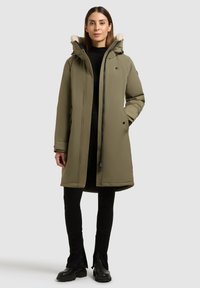 Olivengrøn polstret parka med pelsforet hætte, lynlås, sidelommer og en afslappet pasform, parret med sorte leggings og støvler.