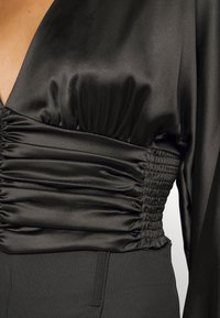 Top en satin noir avec détail de taille élastique froncée, manches longues et décolleté en V profond, porté avec un pantalon noir taille haute.