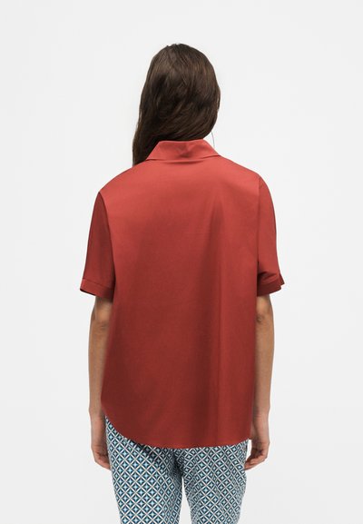 Femme aux longs cheveux bruns portant une chemise rouge ample à manches courtes et un pantalon bleu et blanc à motifs, debout de dos sur un fond blanc.