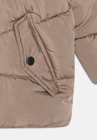 Veste matelassée en tissu beige avec une texture quiltée, dotée d'une poche latérale avec un bouton-pression noir et un éclat subtil.